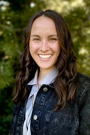 Kaiti Freeberg smiling in a jeans jacket and lavender top in front of a forested setting